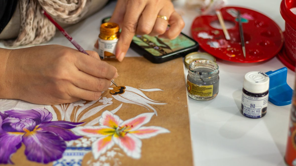 Artist painting vibrant flowers on paper using gouache paints in an indoor setting.